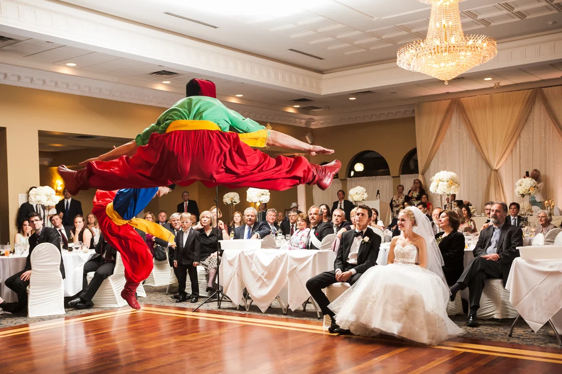 A dancer in red, green and yellow costume in a mid-air split leap on a ballroom dance floor in front of a seated wedding party