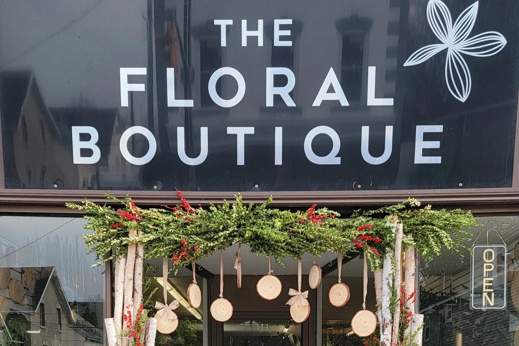 The same sign in winter, with a garland of cedar and red berries strung under it and round wood-slice ornaments hanging from the greenery; a neon OPEN sign glows in the window.