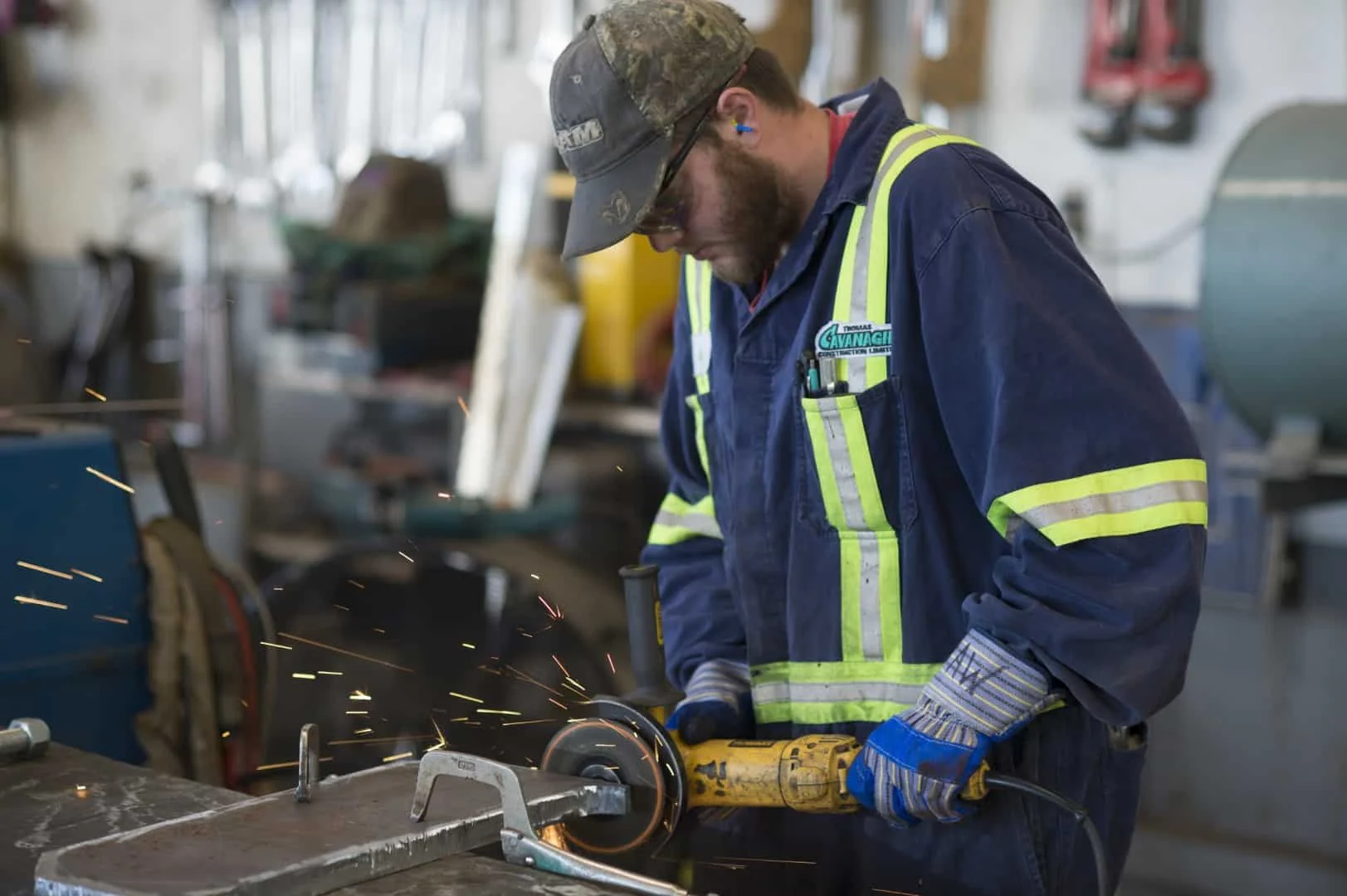 A tradesperson in Cavanagh coveralls and high-visibility stripes cutting steel with an angle grinder in the shop, sparks flying