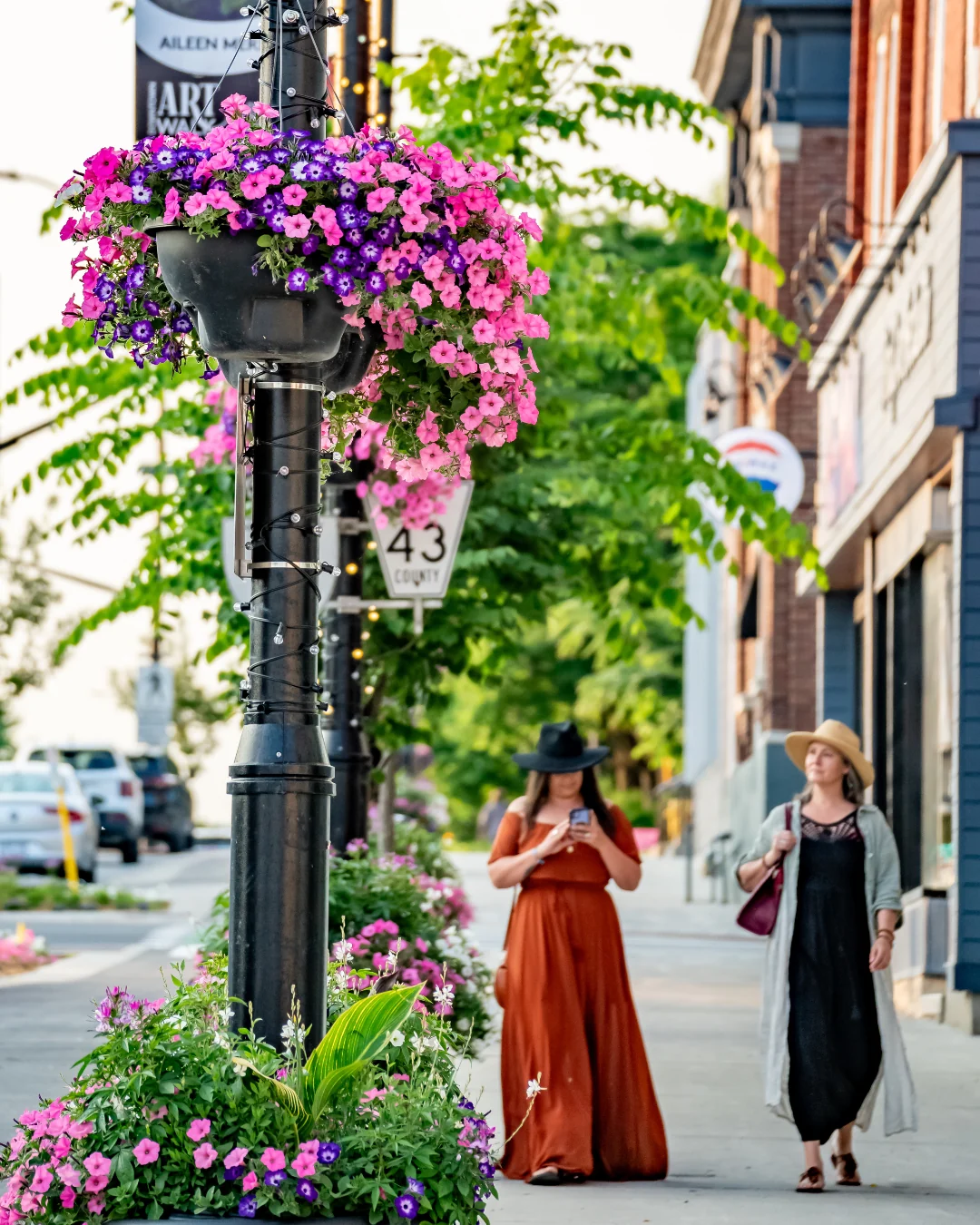 Beckwith Street in summer: a black lamp post with a hanging basket of pink and purple petunias, County Road 43 sign, brick storefronts and two people walking the sidewalk