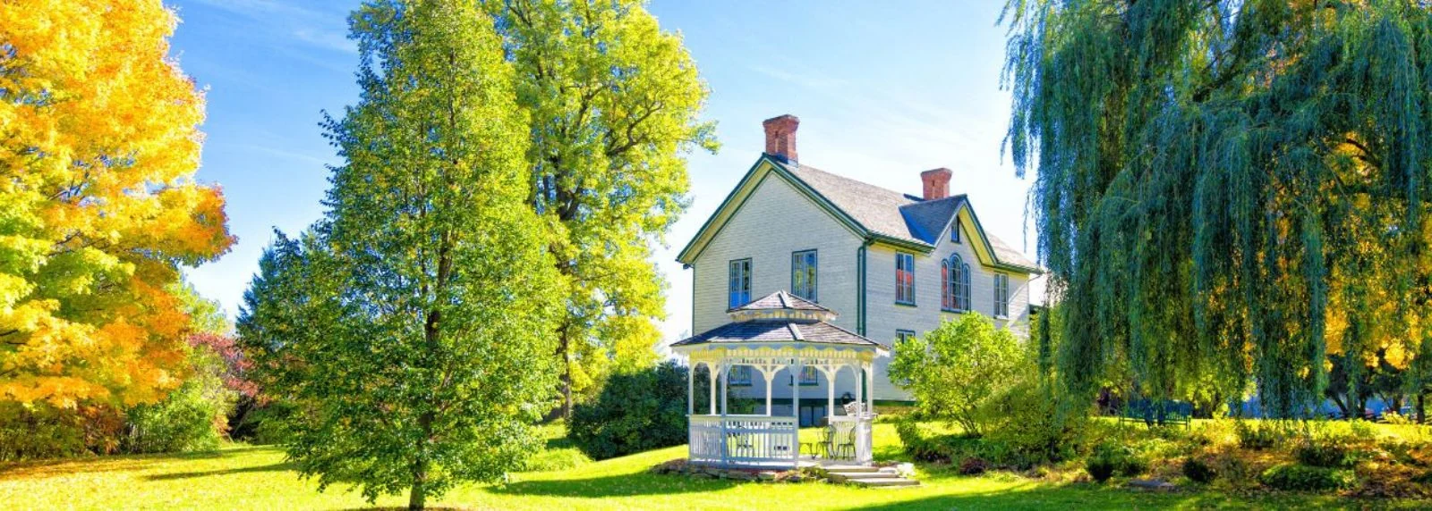 Heritage House Museum - the pale 1861 house behind a white gazebo, a weeping willow to the right and maples turning yellow to the left, on a green lawn
