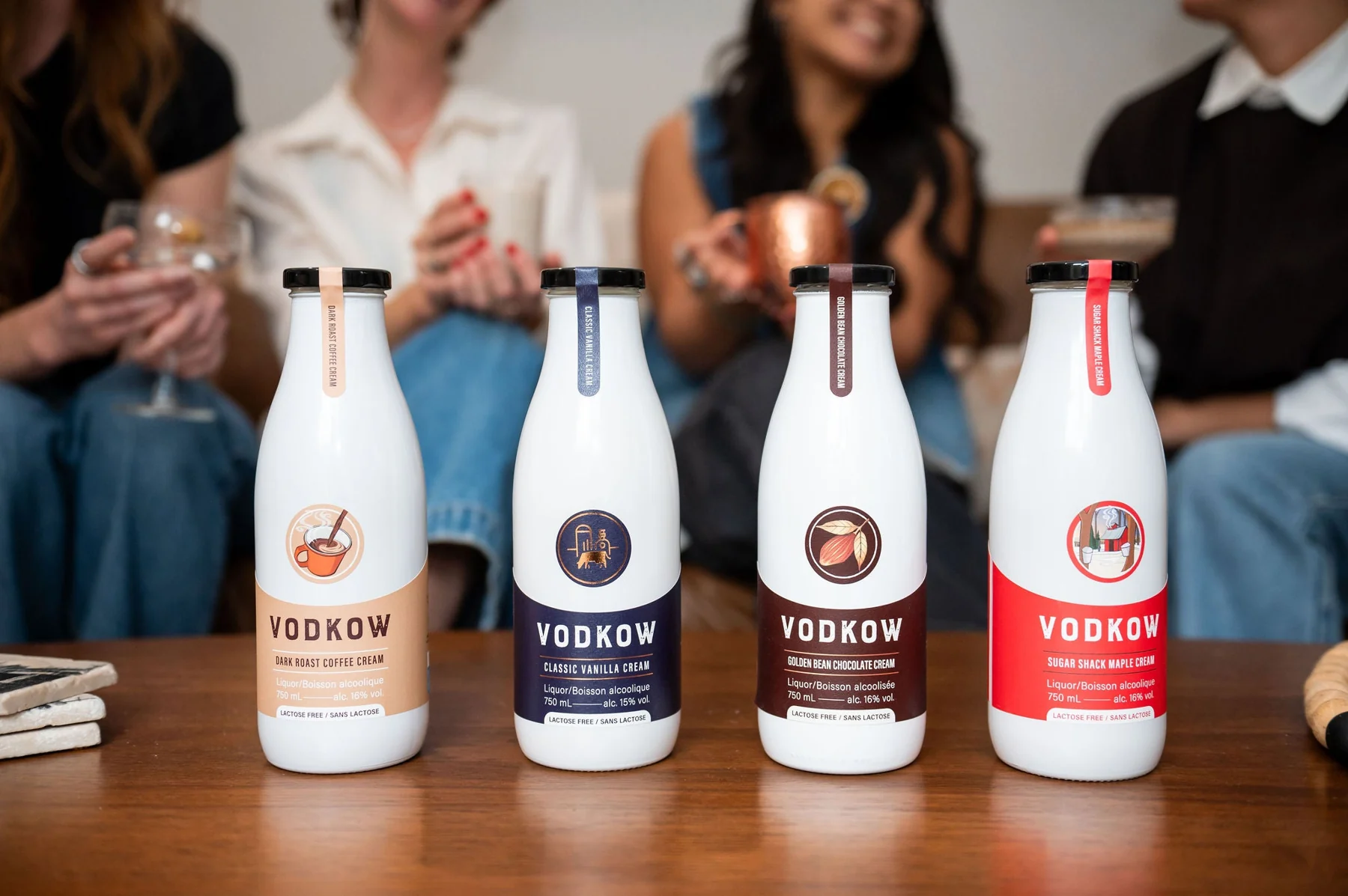 Four Vodkow cream liquor bottles standing on a wooden table, each marked lactose free, with people out of focus holding drinks behind them