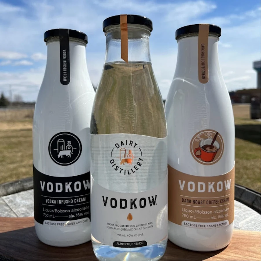 Three Vodkow bottles outdoors on a barrel — Vodka Infused Cream, the Dairy Distillery vodka and Dark Roast Coffee Cream — with a field and open sky behind