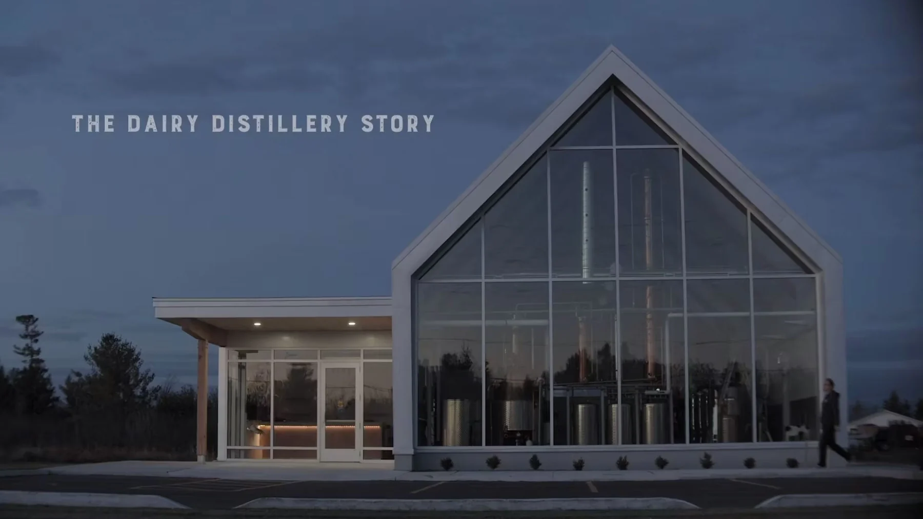 The distillery building at dusk, its tall glass gable showing the fermentation tanks and copper still inside, with the caption "The Dairy Distillery Story"